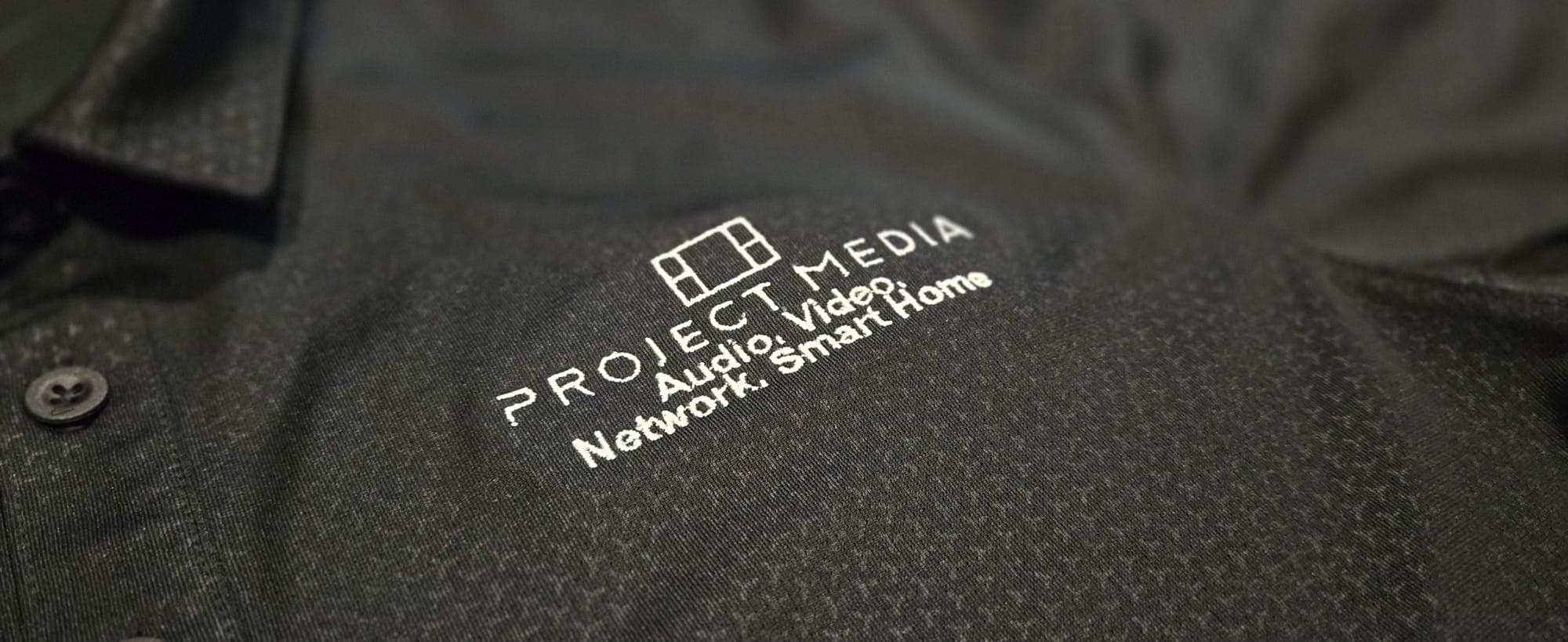 Close-up of a branded Project Media company polo shirt with the logo and Audio, Video, Network, Smart Home tagline
